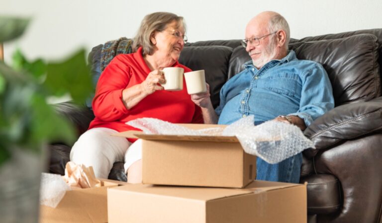Senior couple sitting on a sofa, toasting mugs while unpacking boxes with packing paper nearby.