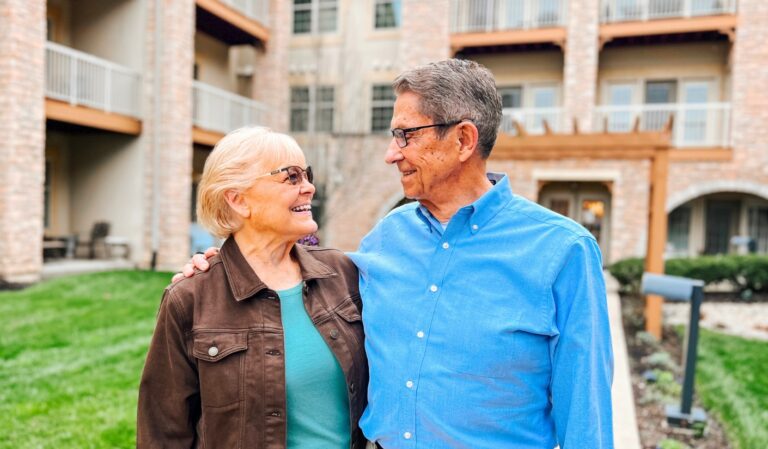 Elderly couple smiling at each other outside an apartment building, with arms around each other.
