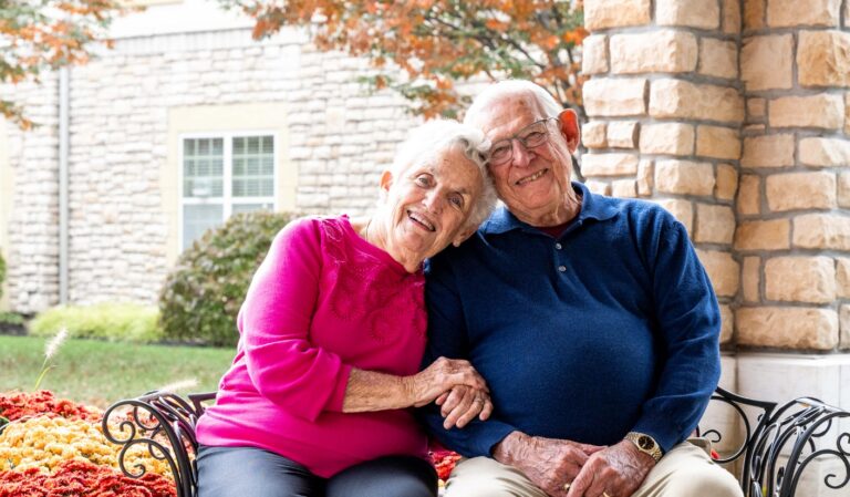 Smiling elderly couple sits on a bench outside a stone building, holding hands.