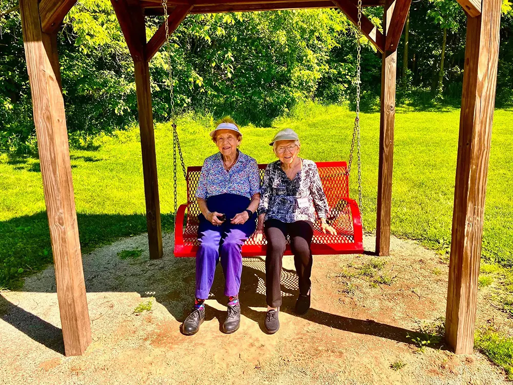 Two elderly women sit together on a red swing bench under a wooden gazebo in a sunny park.