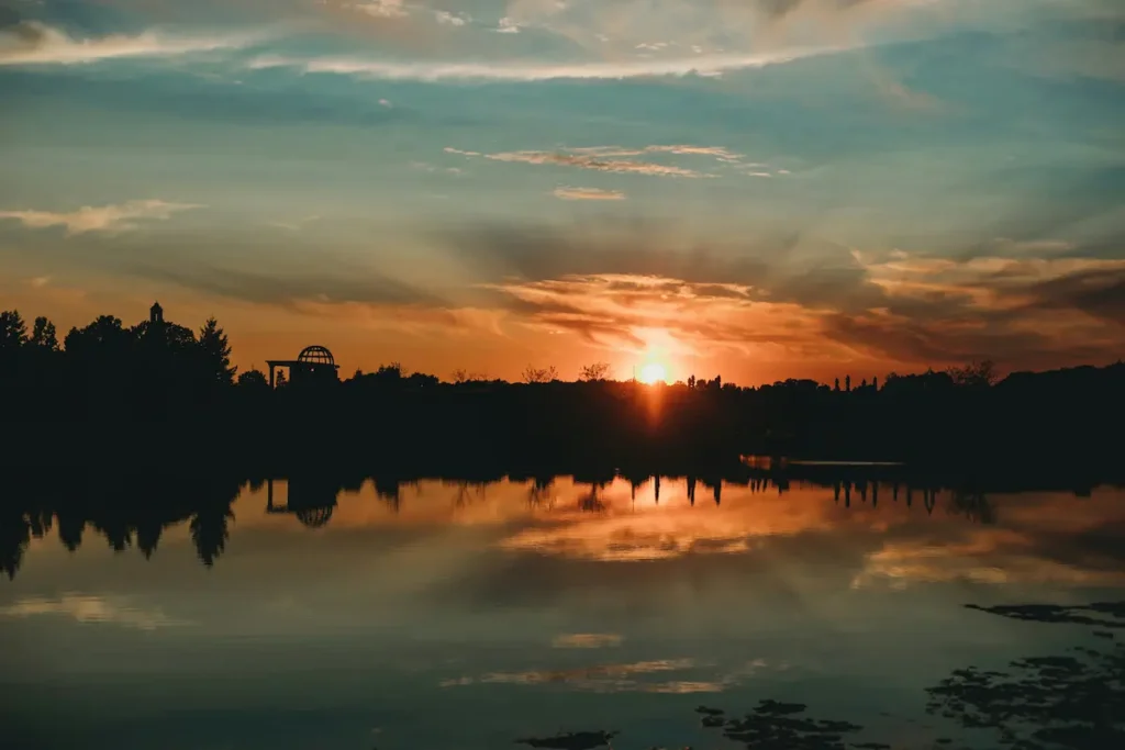 Sun setting over a calm lake with silhouetted trees and a small domed pavilion reflected in the water in a warm orange glow?