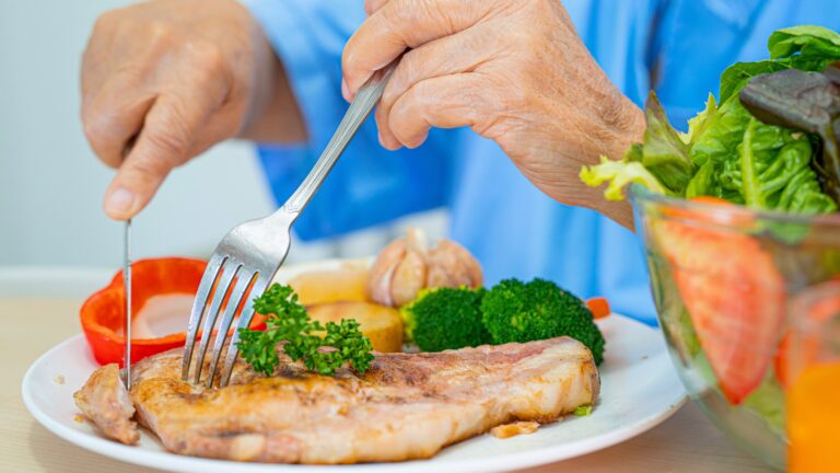 Close-up of hands cutting a cooked chicken breast with a fork and knife on a plate surrounded by broccoli and herbs.