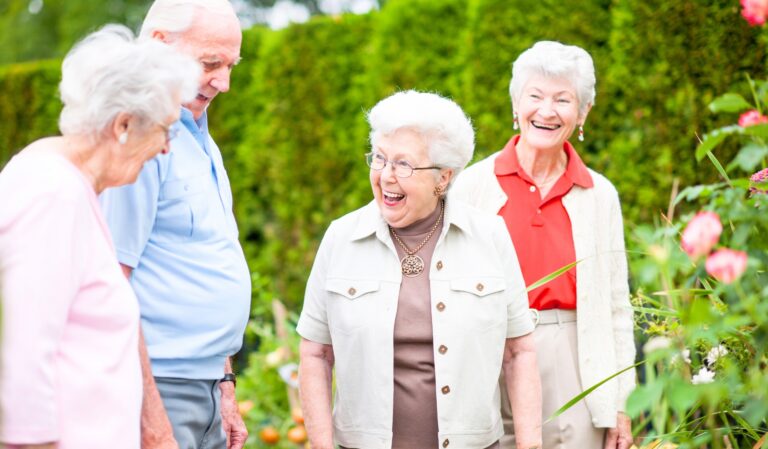 Four senior friends stand in a garden, smiling and chatting among flowers and greenery.